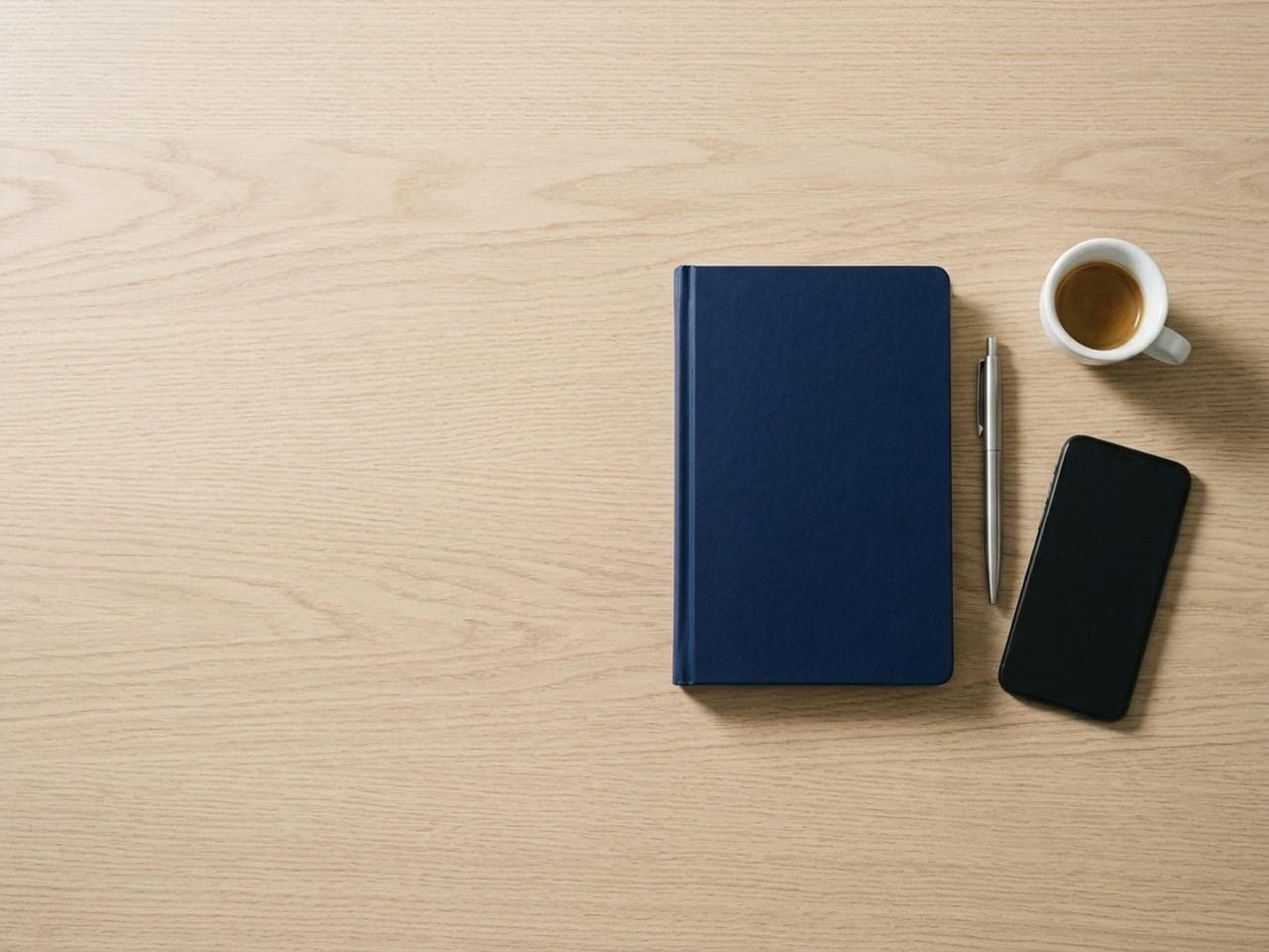 Navy notebook, pen, phone and coffee cup on a pale oak desk